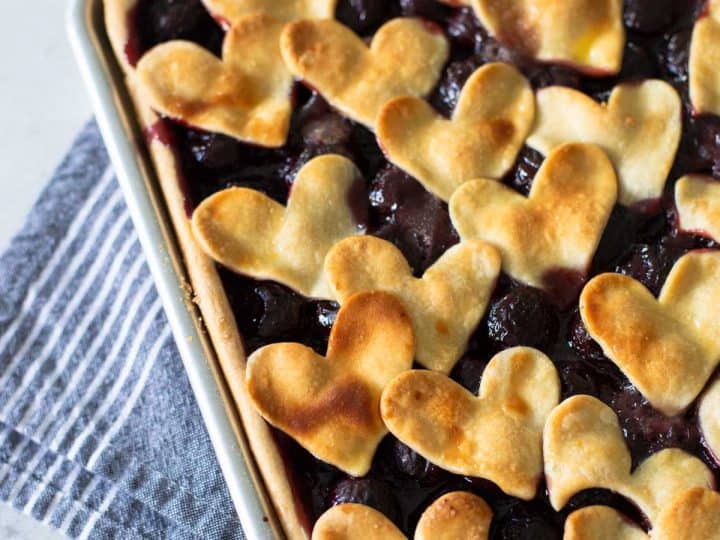 A cherry slab pie sits in a metal baking pan and has heart-shaped cut outs for the top crust.