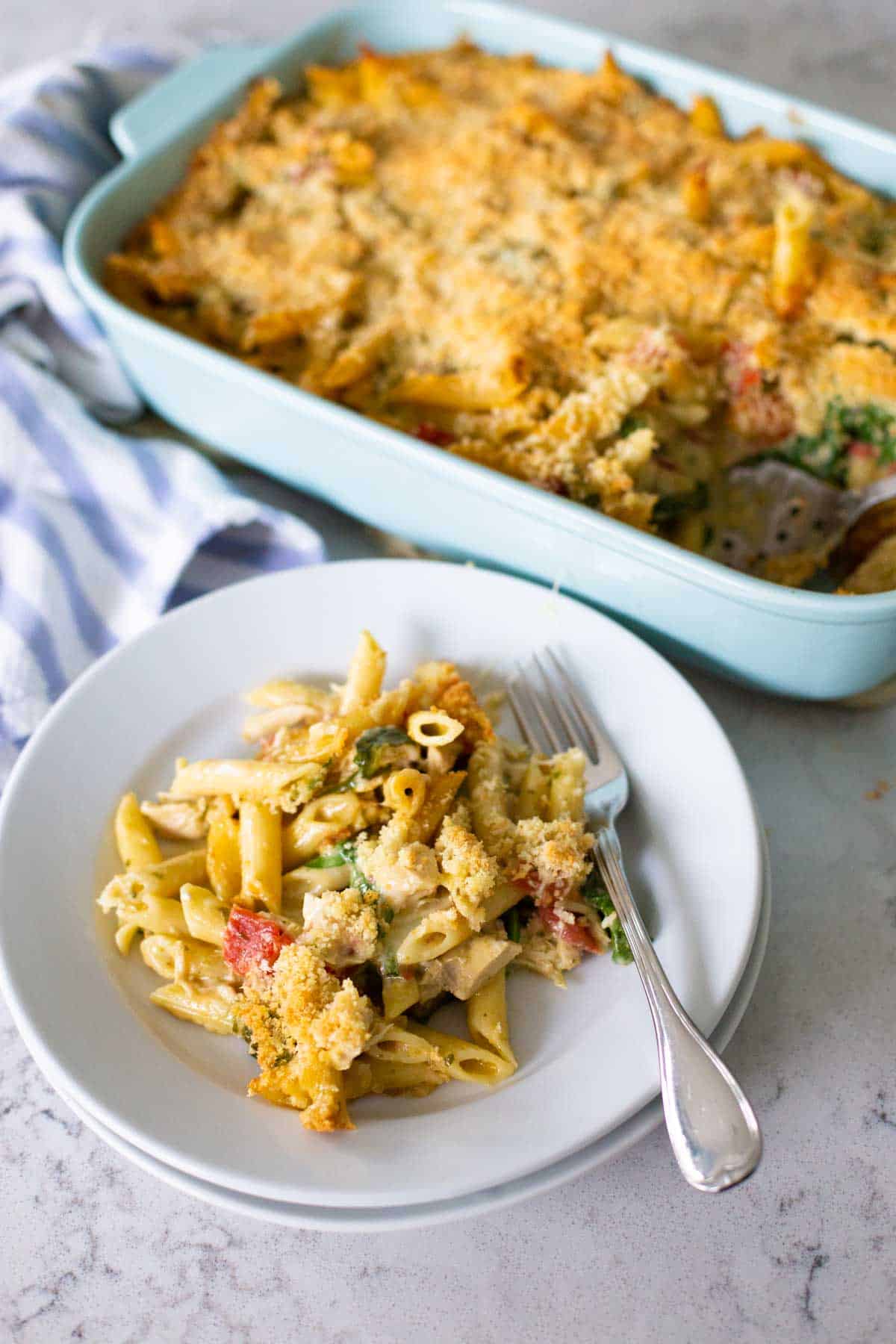 The baking dish is next to a white plate with a serving of the chicken pasta casserole.