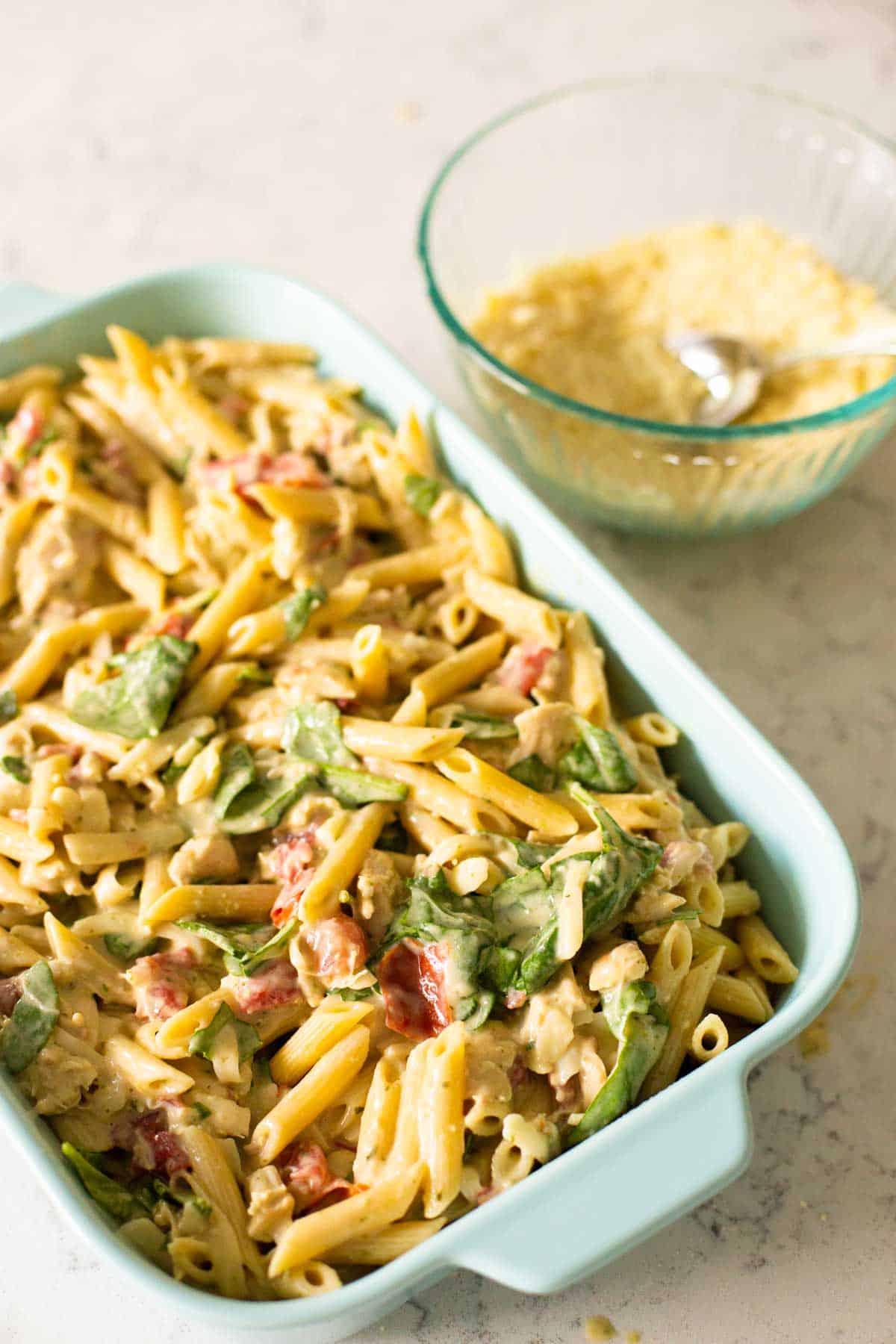 The chicken pesto casserole is in a blue baking dish ready to be topped with bread crumbs.