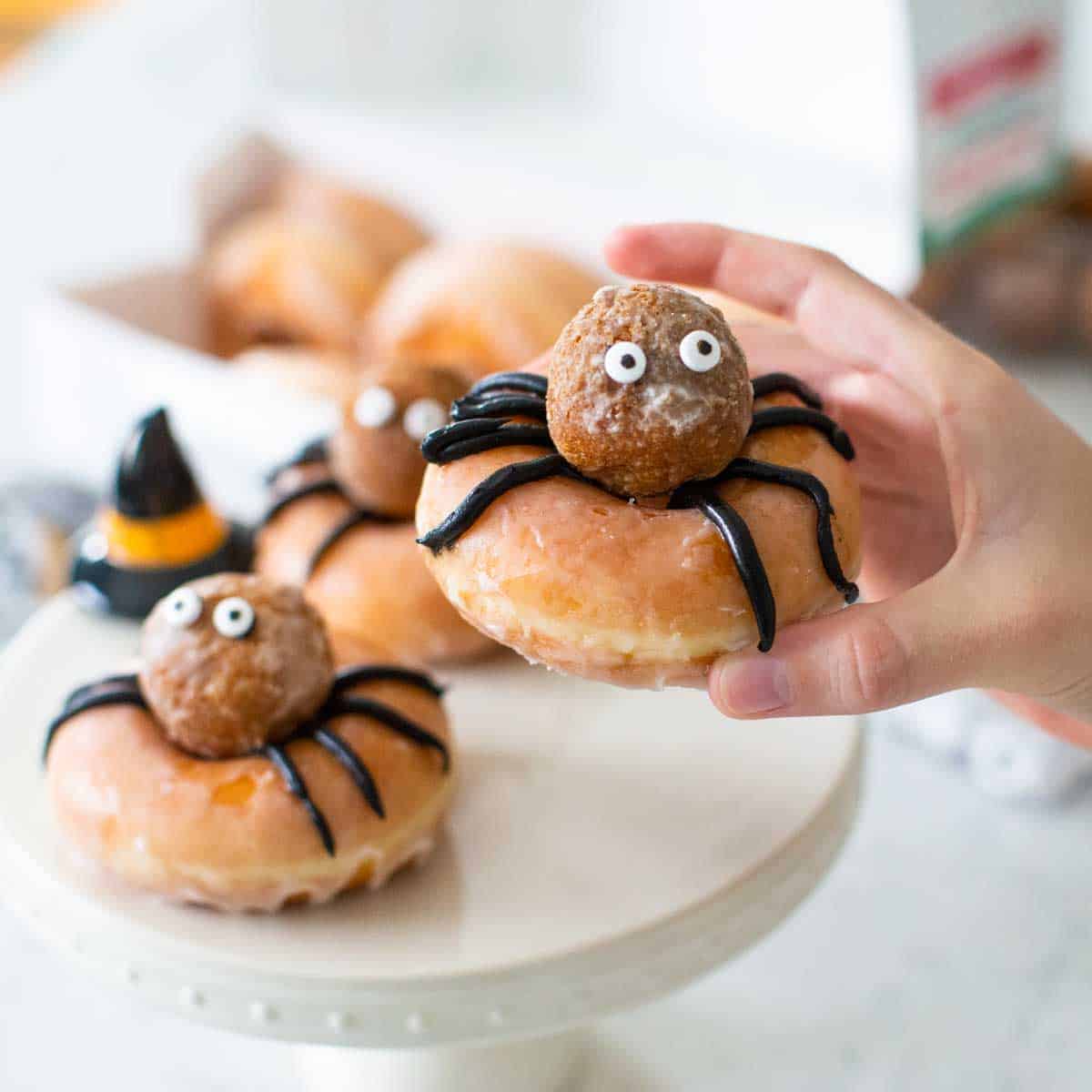 A glazed donut decorated to look like a spider with a donut hole on top, googly eyes, and black frosting legs is in the hand of a kid.