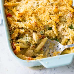 A blue baking dish filled with alfredo pesto pasta and topped with crunchy breadcrumbs. A slotted spoon has scooped out a serving.