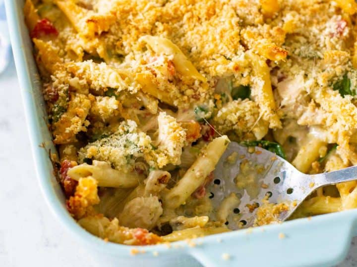 A blue baking dish filled with alfredo pesto pasta and topped with crunchy breadcrumbs. A slotted spoon has scooped out a serving.