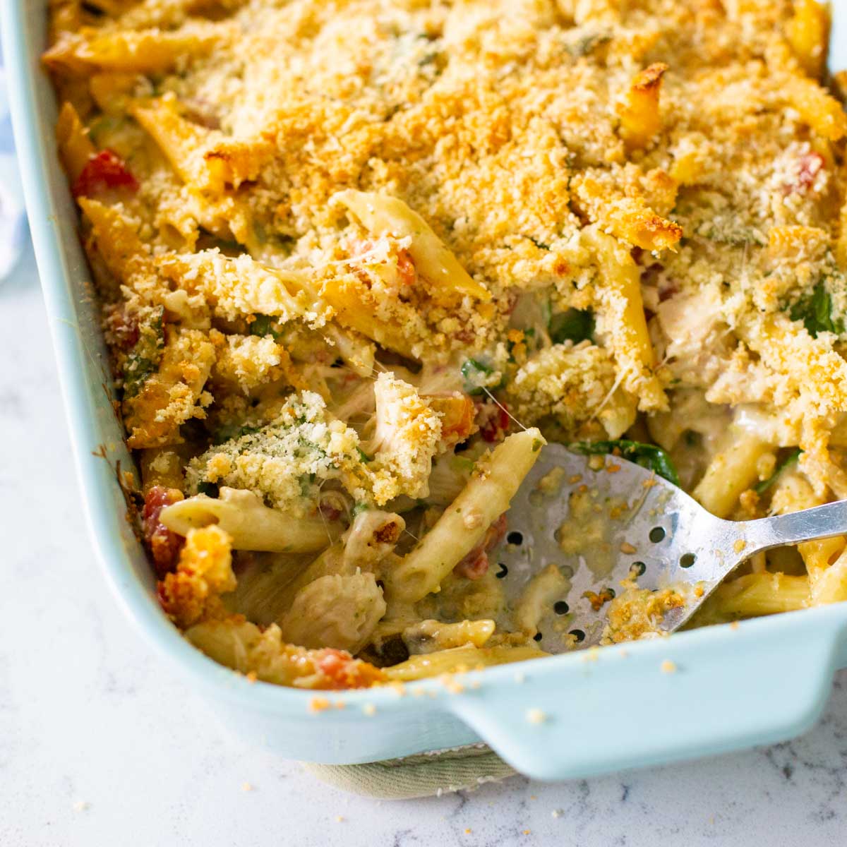 A blue baking dish filled with alfredo pesto pasta and topped with crunchy breadcrumbs. A slotted spoon has scooped out a serving.