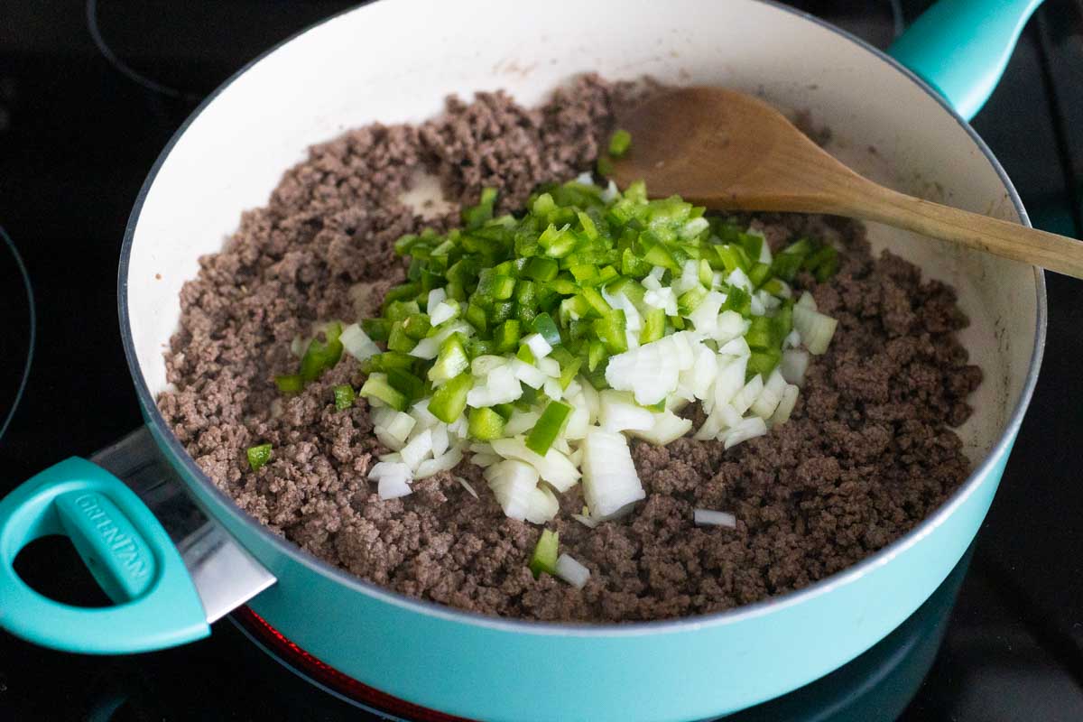 The blue skillet has the browned ground beef with chopped peppers and onions on top. A wooden spoon is about to stir them together.