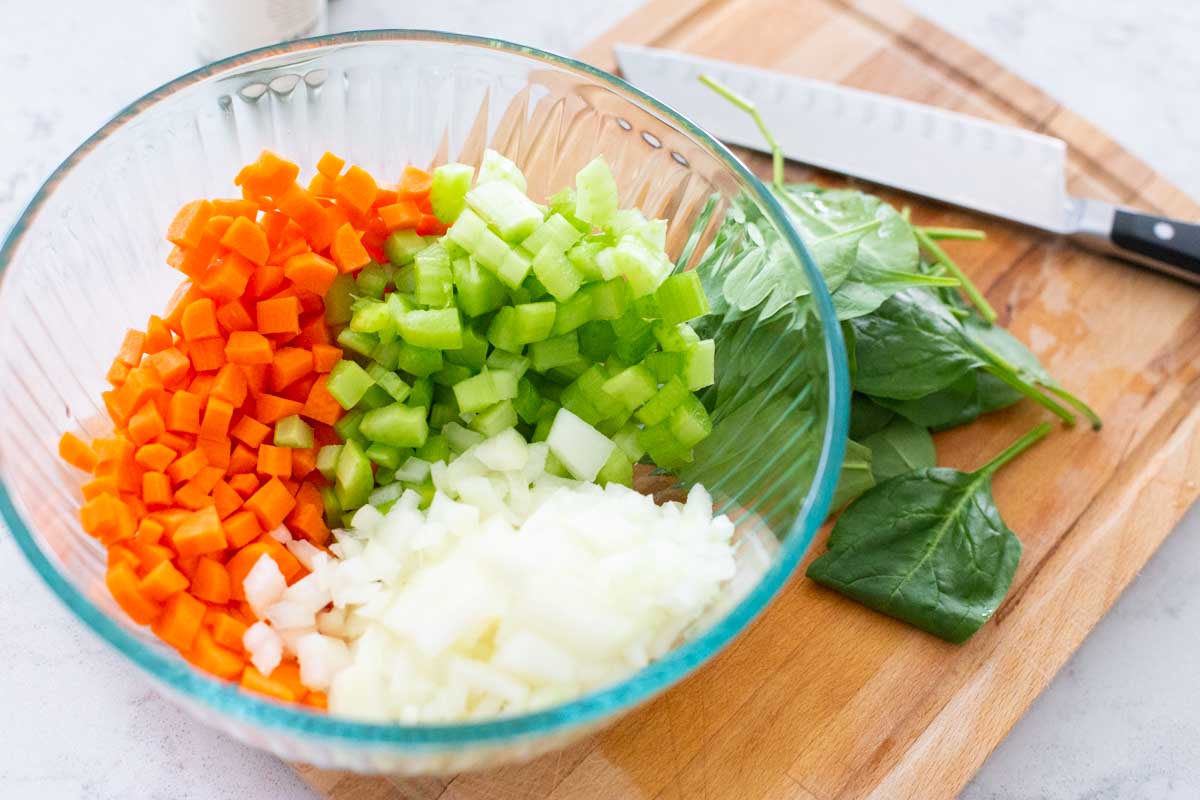 The diced onion, carrots, and celery are in a large mixing bowl next to a cutting board with a chef knife.