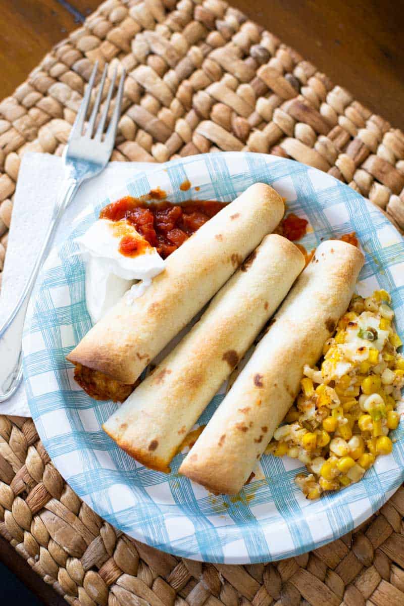 A blue and white dinner plate has 3 taquitos, corn, sour cream, and salsa.