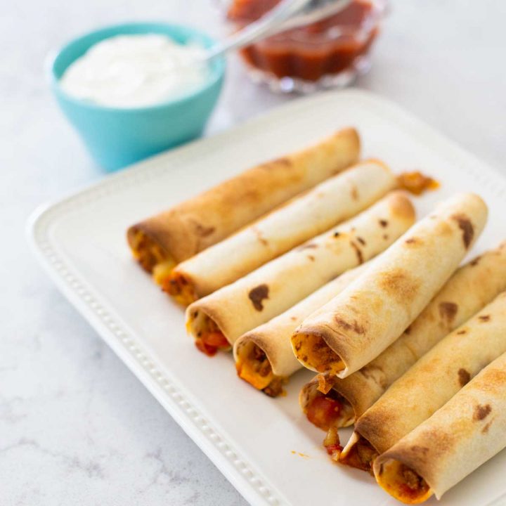 A platter of rolled-up turkey taquitos sits next to a bowl of sour cream and a bowl of salsa.