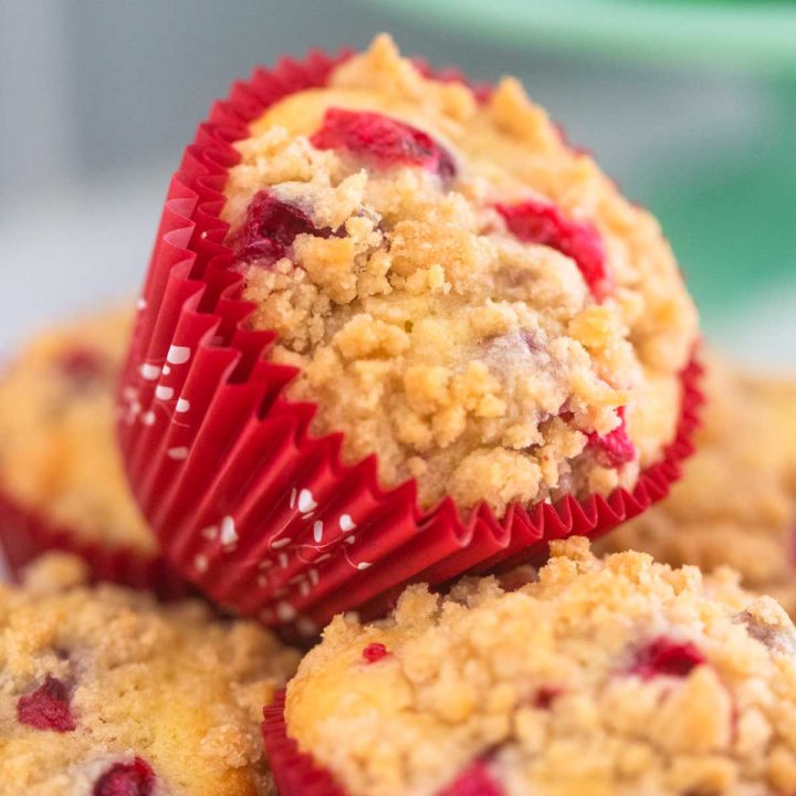 A cranberry orange muffin with cinnamon streusel is wrapped in a bright red muffin paper and sits on a pile of other muffins.
