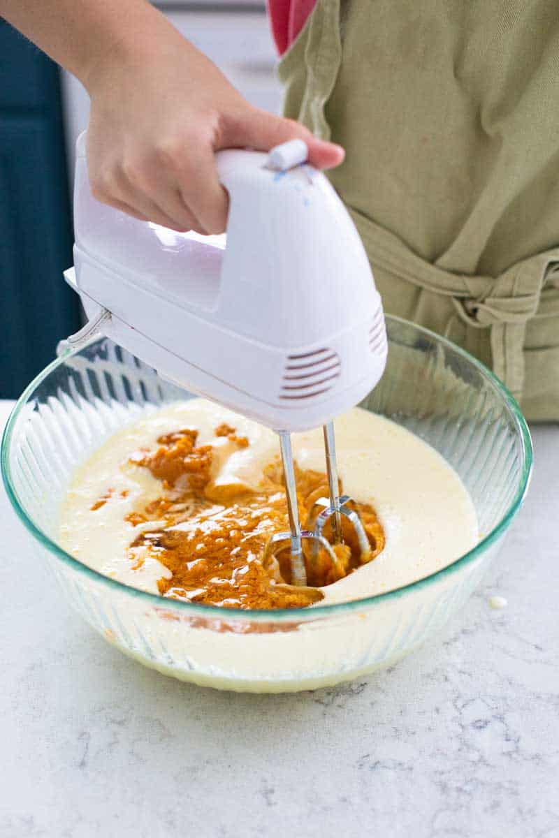 A girl uses a hand mixer to beat a bowl of pumpkin cookie batter.