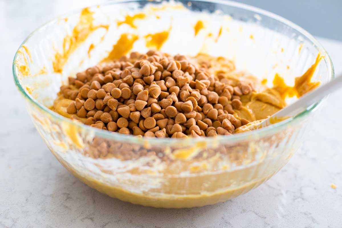A big bowl of pumpkin cookie batter has butterscotch chips on top ready to be stirred in.