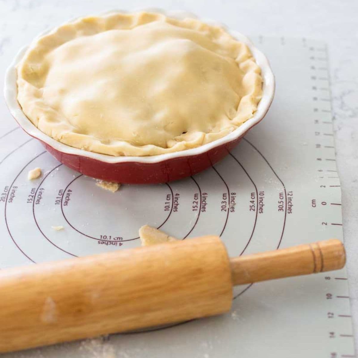 The apple pie has been assembled in the dish and sits on the rolling mat next to the rolling pin.
