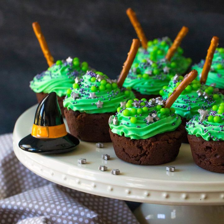 A cake plate has several brownie bites with green frosting and pretzel rods that look like witch cauldrons. There is a black witch hat decoration on the plate.