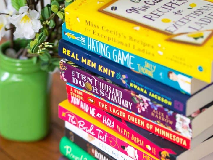 A stack of colorful book club fiction books next to a green vase of flowers.