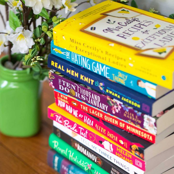 A stack of colorful book club fiction books next to a green vase of flowers.