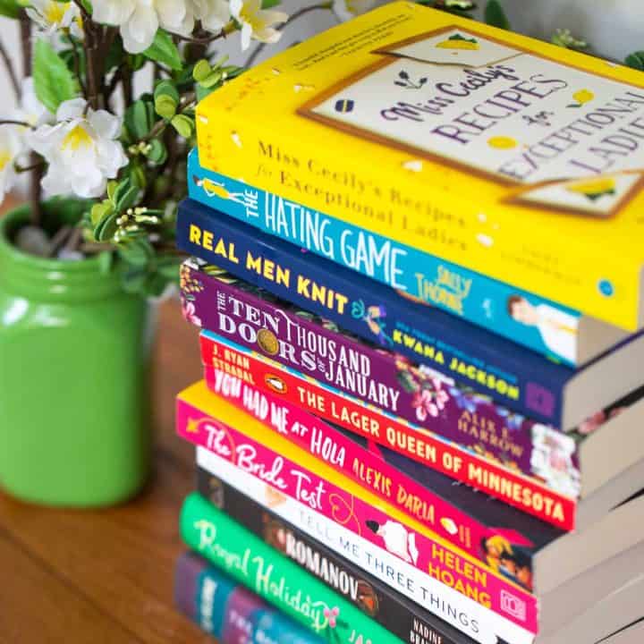 A stack of colorful book club fiction books next to a green vase of flowers.