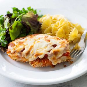 A dinner plate has a serving of baked chicken parmesan, farfalle pasta, and a green salad.