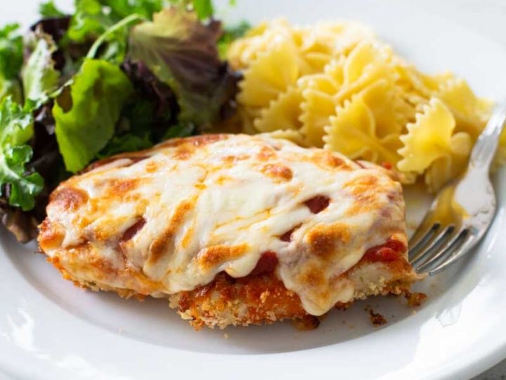 A dinner plate has a serving of baked chicken parmesan, farfalle pasta, and a green salad.