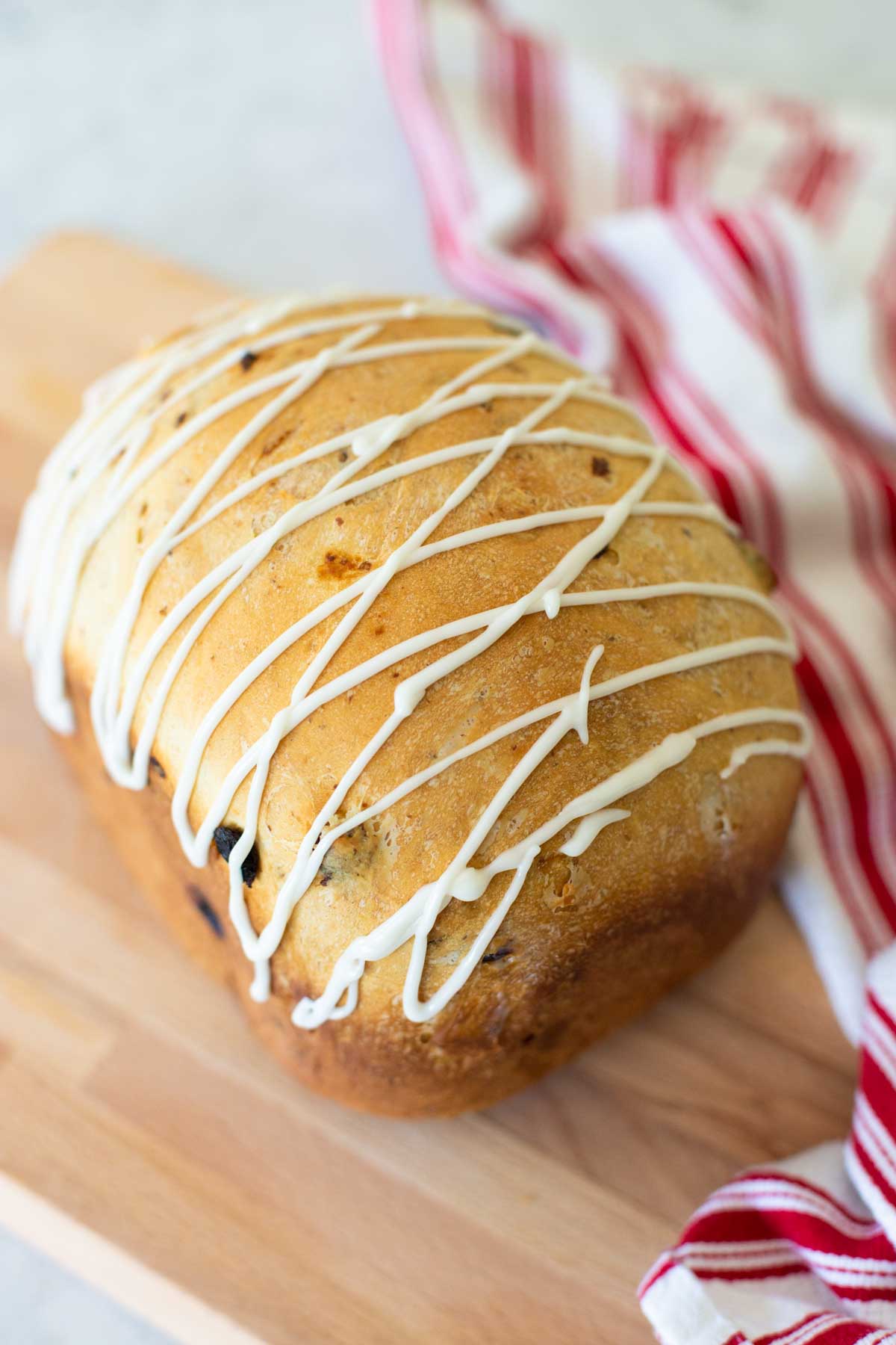 A finished loaf of Bavarian Christmas bread from the bread machine. The loaf has a drizzle of white glaze in a criss cross pattern over the top and a red and white striped towel sits next to it.