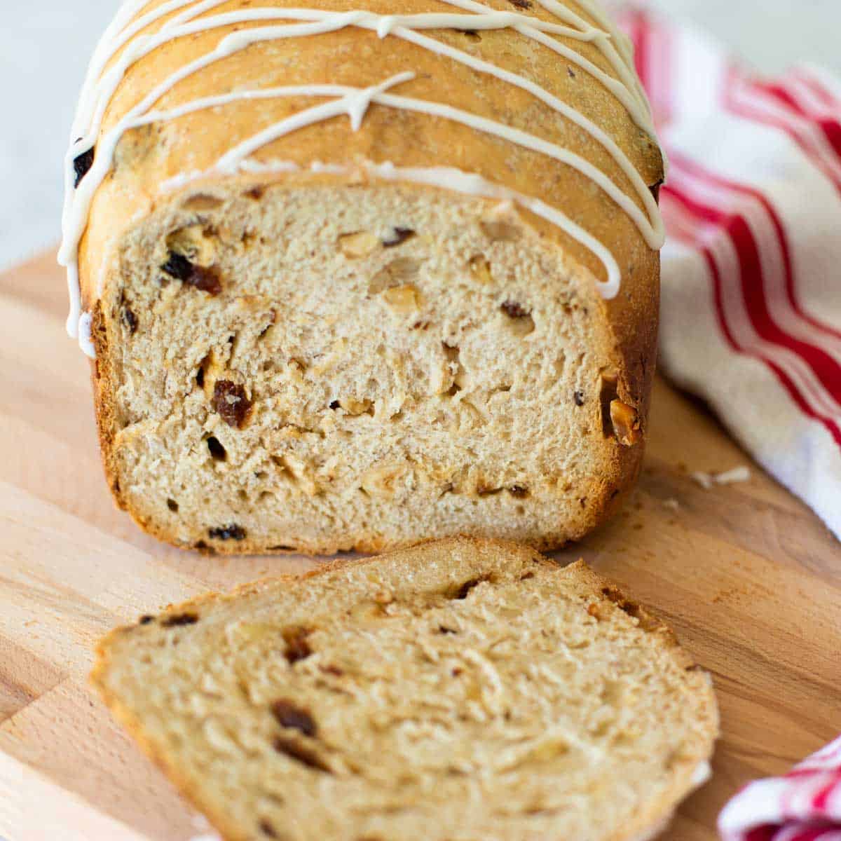 A finished loaf of Bavarian Christmas bread from the bread machine. The loaf has a drizzle of white glaze in a criss cross pattern over the top and a red and white striped towel sits next to it.