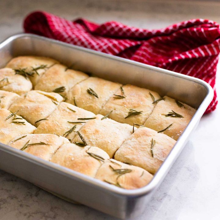 A metal baking pan filled with square-shaped rosemary dinner rolls with roasted herbs over the top.