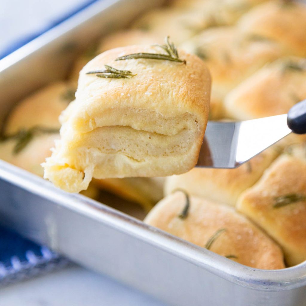 A spatula is lifting a rosemary pull apart roll from the 9x13-inch baking pan it was baked in.