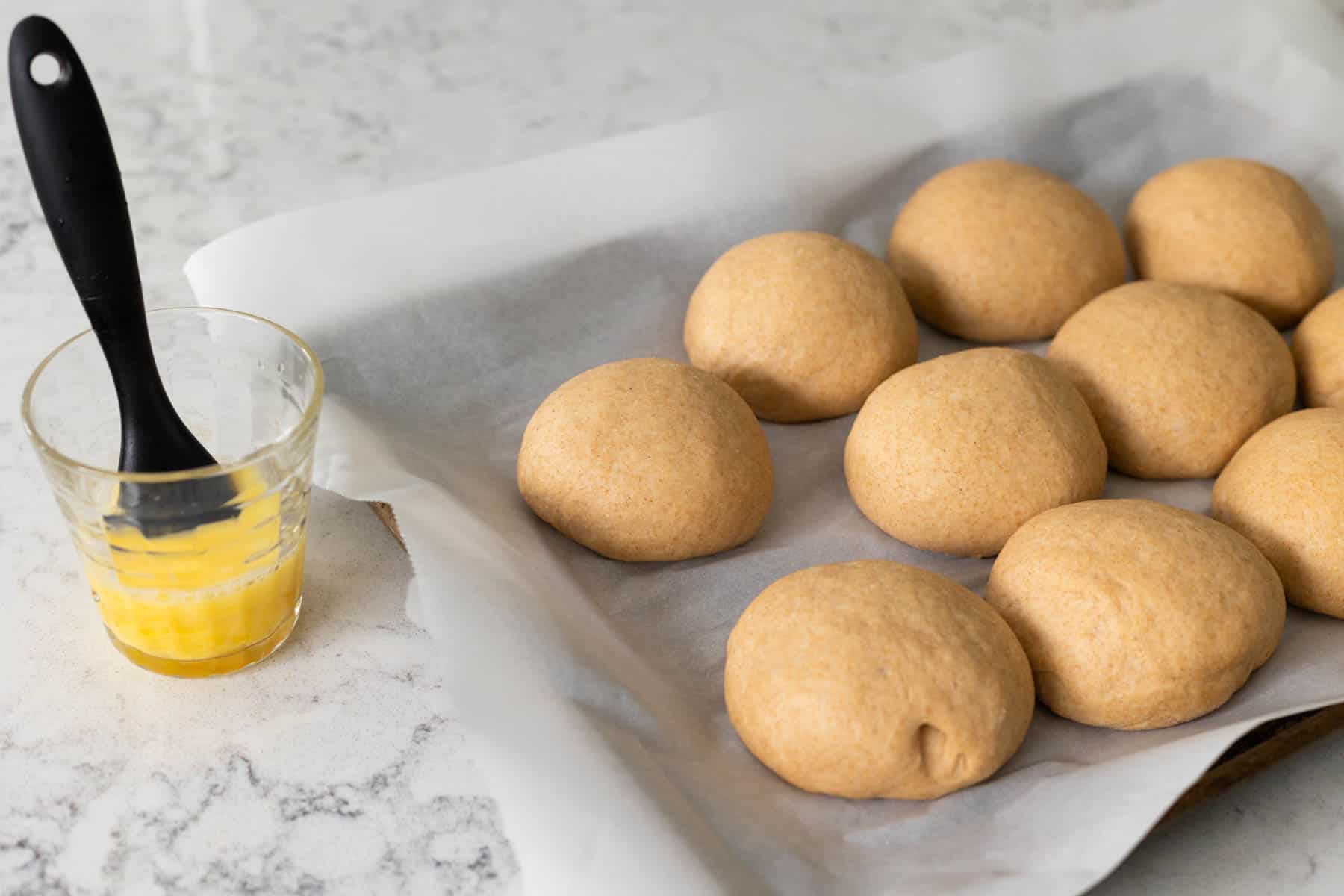 A cup of egg wash has a pastry brush in it next too the pan of risen rolls.