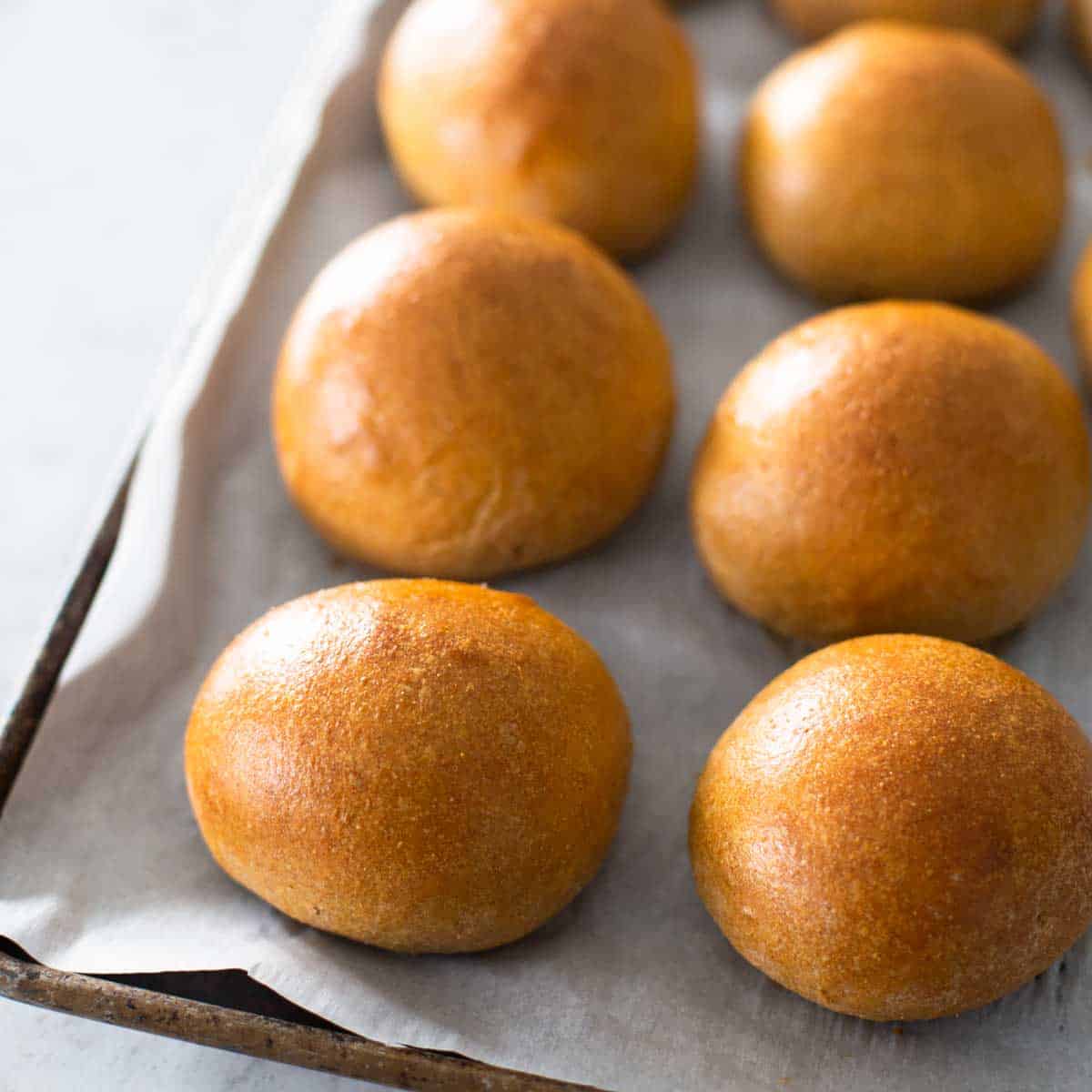 A pan of golden brown whole wheat hamburger buns fresh from the oven. The pan is lined with parchment paper.