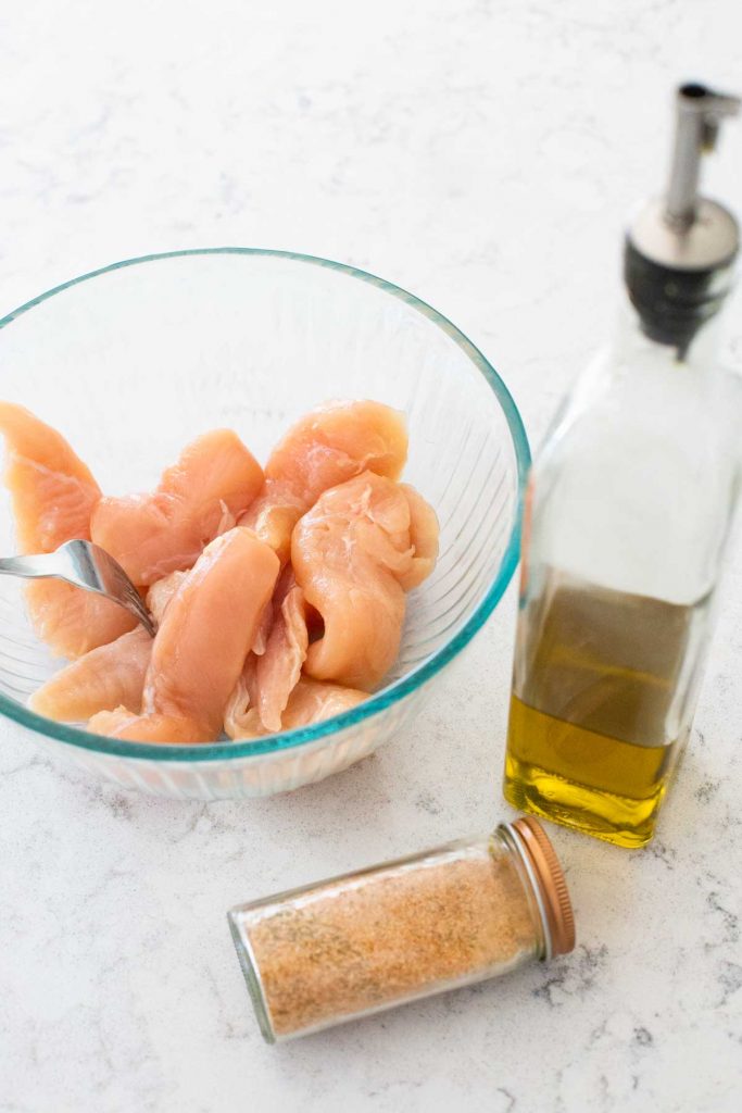 A bowl of raw chicken tenderloins sits next to a bottle of olive oil and a jar of seasoning.