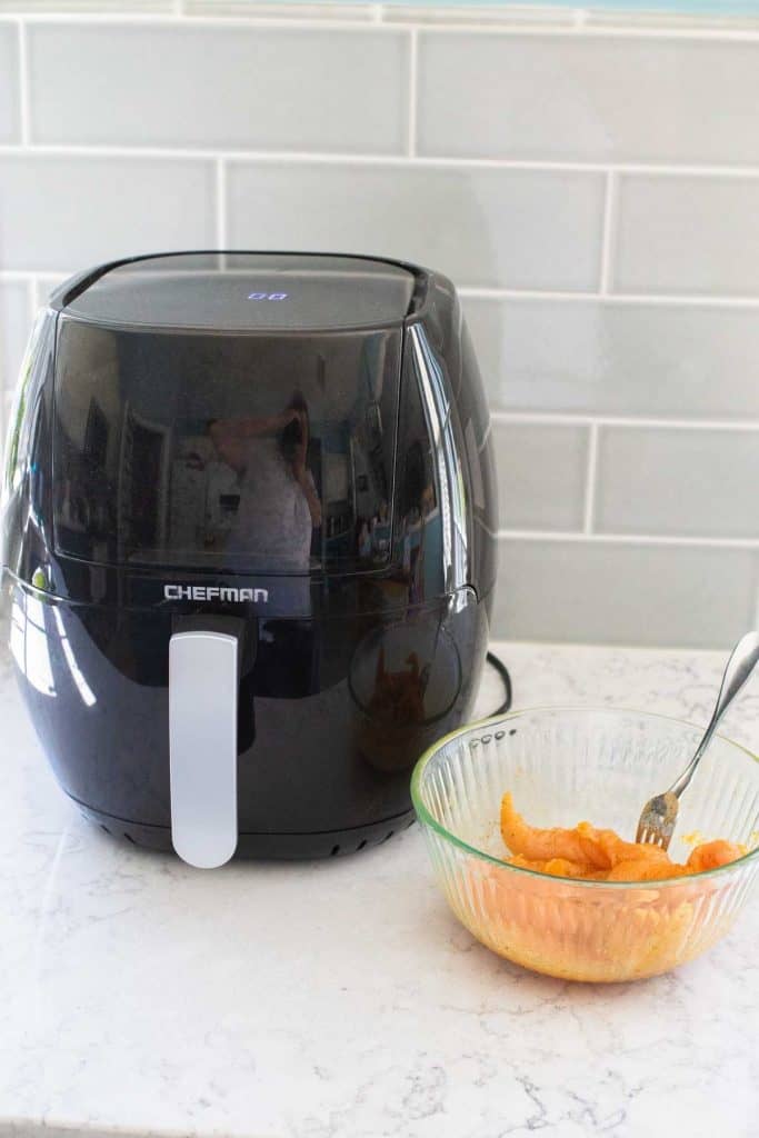 A black airfryer sits on a kitchen counter next to a bowl of seasoned chicken tenderloins.