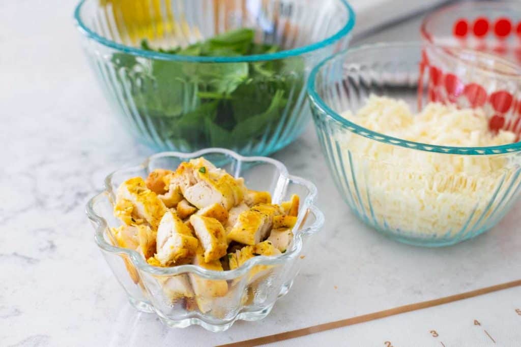 A bowl of chopped chicken tenderloins sits next to other prep bowls filled with vegetables and shredded cheese.