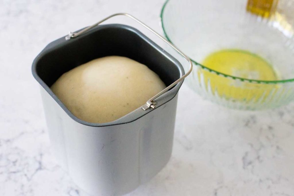 Risen pizza dough in a bread machine bread pan sits next to a bowl brushed with olive oil.