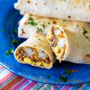 A blue plate has a stack of breakfast burritos, one is sliced open to show the sausage, eggs, and potatoes inside.