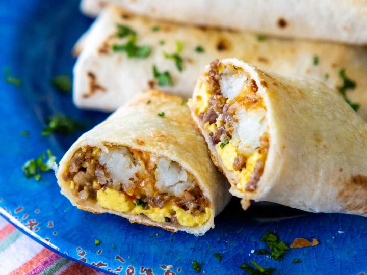 A blue plate has a stack of breakfast burritos, one is sliced open to show the sausage, eggs, and potatoes inside.