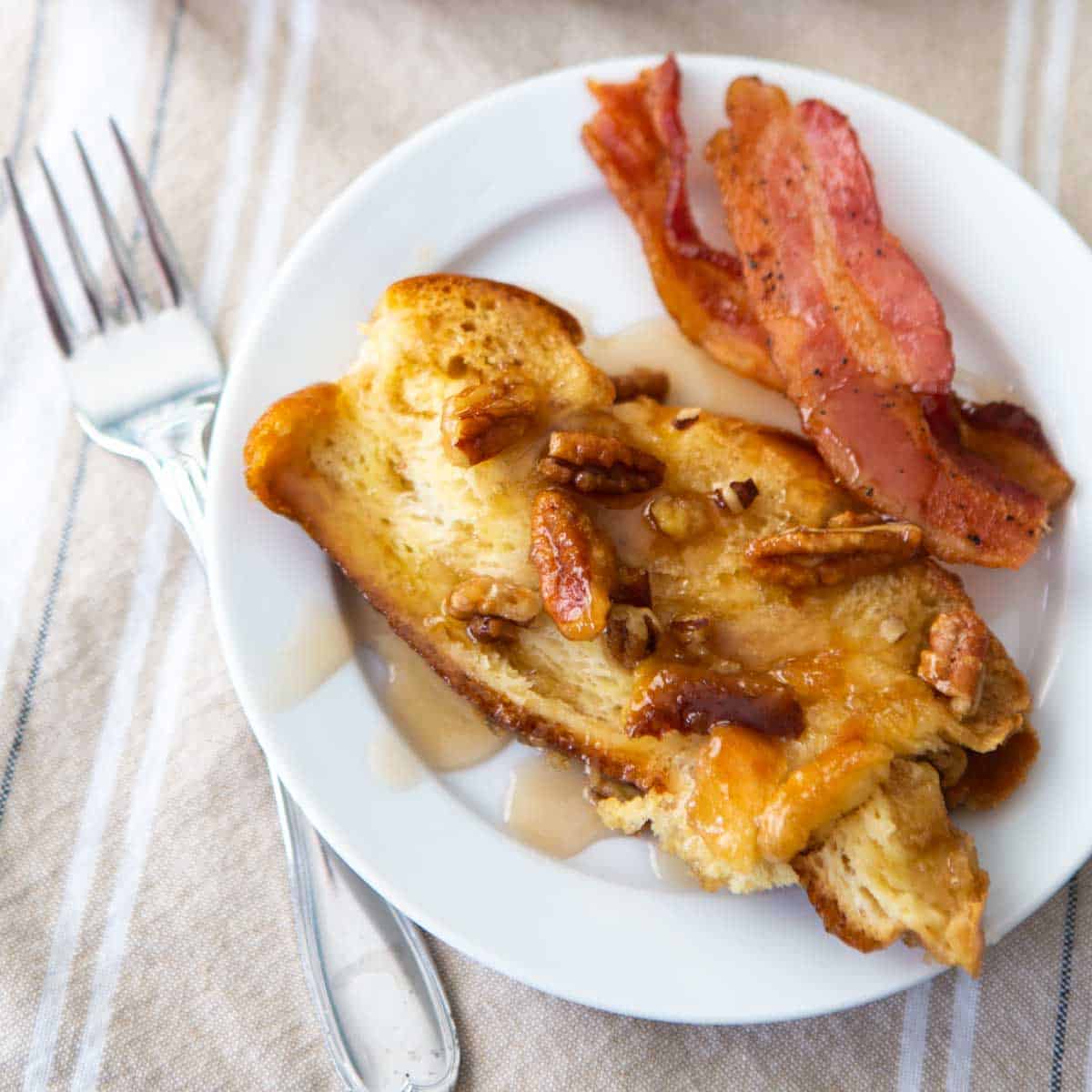 A plate with bourbon maple french toast has pecans and bacon next to it. Maple syrup is drizzled over the top.