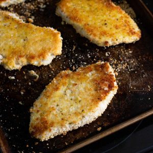 3 chicken breasts coated in bread crumbs have been baked in the oven on a dark baking sheet to get a golden brown crust.