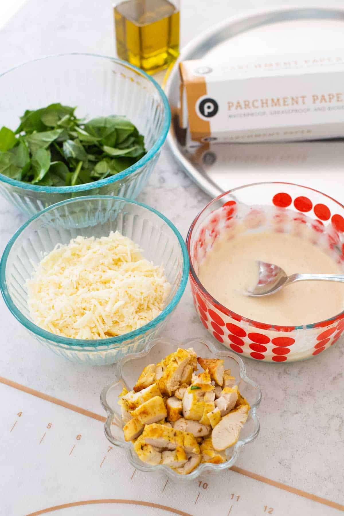 The ingredients for a homemade chicken spinach alfredo pizza are on the counter: a bowl of spinach, shredded cheese, chopped chicken, and homemade alfredo sauce.