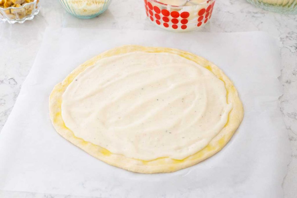 A pizza dough has been rolled out and covered in olive oil and Alfredo sauce.