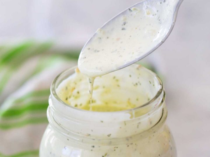 A mason jar with creamy homemade ranch dressing has a spoon showing the texture in a drip.