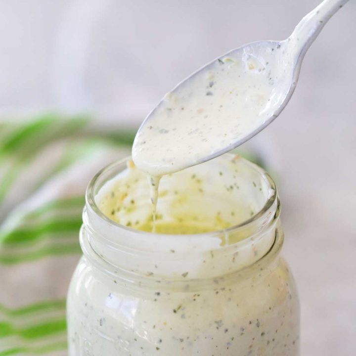 A mason jar with creamy homemade ranch dressing has a spoon showing the texture in a drip.