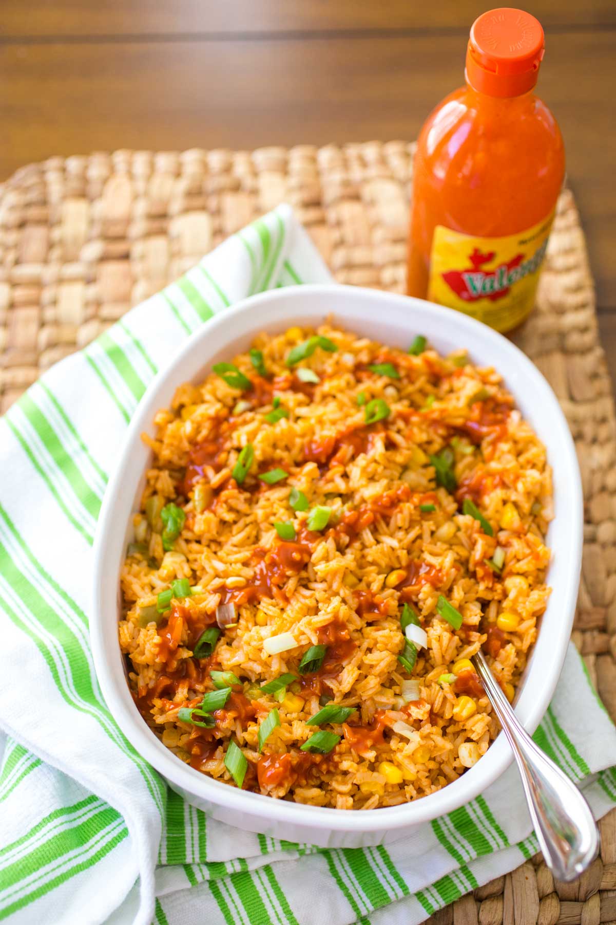 A white baking dish is filled with finished Mexican Rice. Valentina hot sauce is drizzled over the top and chopped green onions are sprinkled for garnish.