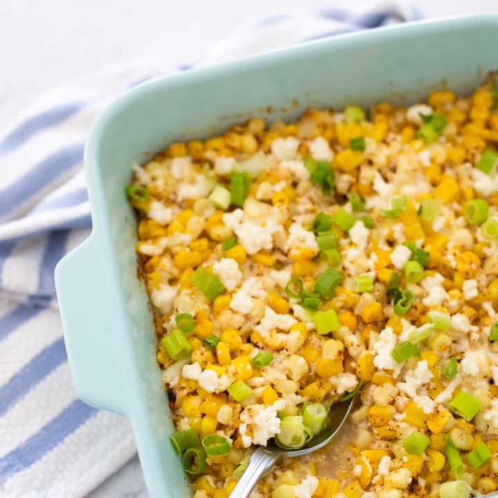 A blue baking dish with yellow Mexican Street Corn casserole has crumbled cheese and fresh green onions sprinkled over the top.