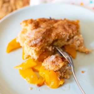 A portion of peach cobbler on a plate with a fork. You can see the juicy fresh peaches under the crust.