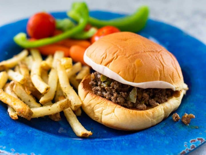 A blue plate has a sloppy joe sandwich next to baked french fries with fresh veggies in the back.