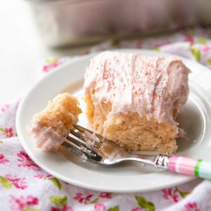 A slice of pink lemonade cake with light pink frosting and sprinkles sits on a white plate. A colorful fork has taken a bite to show the texture of the cake.