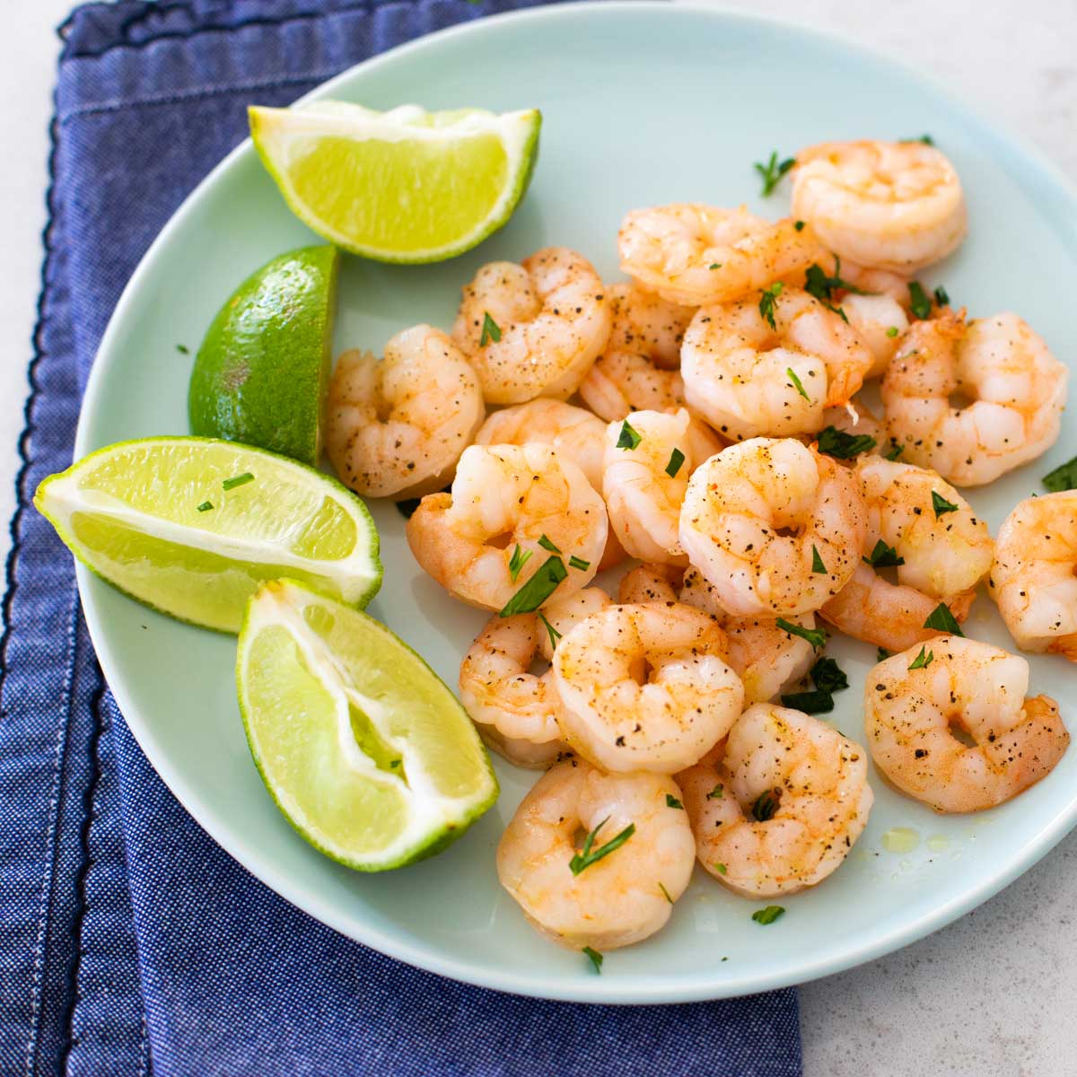 A blue plate with roasted shrimp next to lime wedges. Fresh parsley is sprinkled over the top. A blue napkin is under the plate.