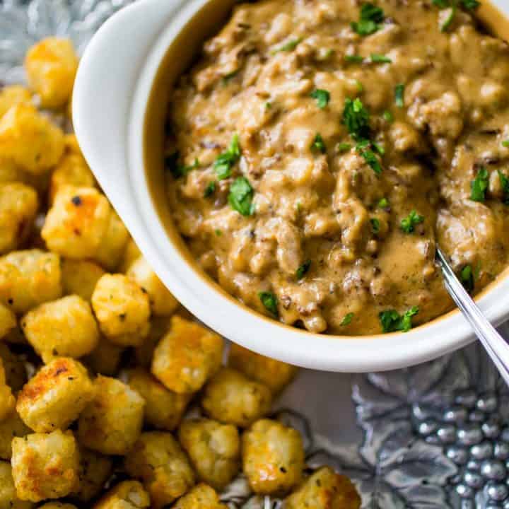 A white bowl filled with hot sausage dip has a serving spoon and a pile of tater tots for dunking on a platter.