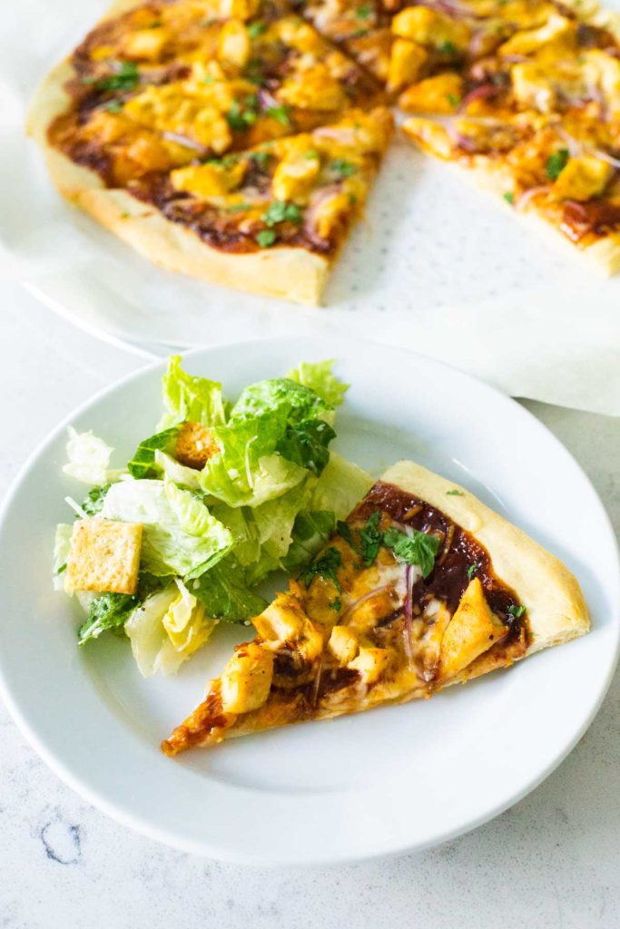 A slice of barbecue chicken pizza sits on a plate next to some caesar salad.