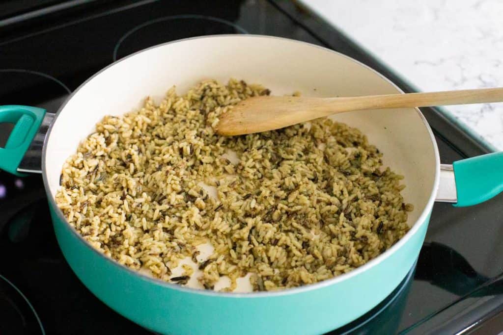 Wild rice is being fried in olive oil in a blue skillet. A wooden spoon is stirring the rice.