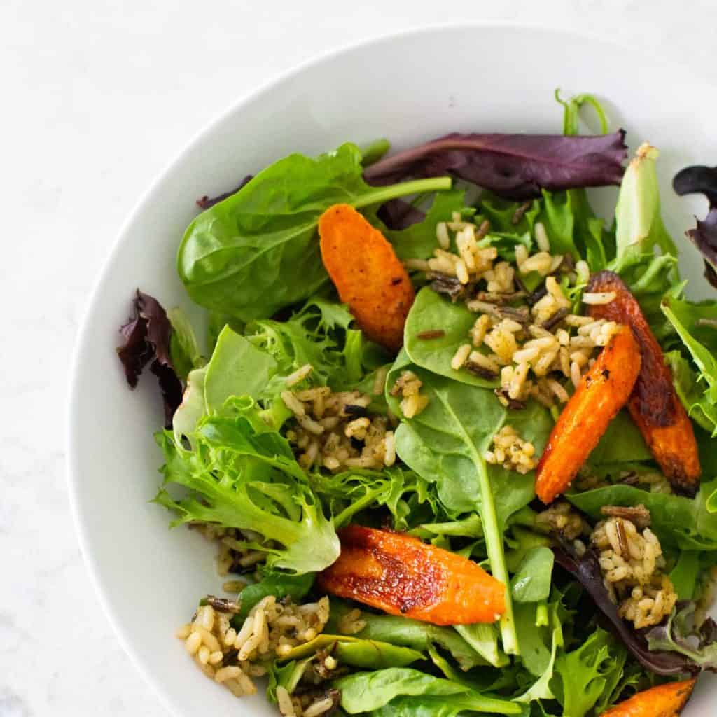 A serving of crispy rice salad shows green lettuce, roasted carrots, fried wild rice in a white bowl.