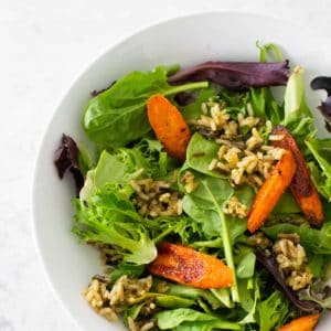 A serving of crispy rice salad shows green lettuce, roasted carrots, fried wild rice in a white bowl.